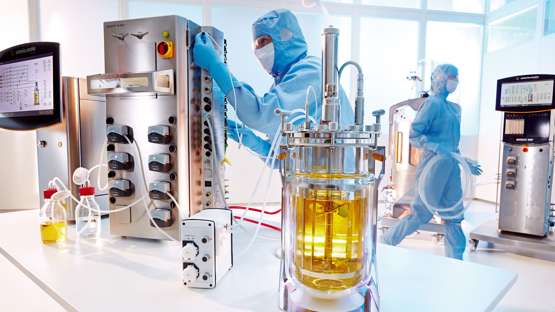 Two people in sterile clothing work in a laboratory with bioreactor and analytical equipment, including a fermentation vessel containing a yellow liquid, while utilizing a biosimulation tool to predict microbial behavior.