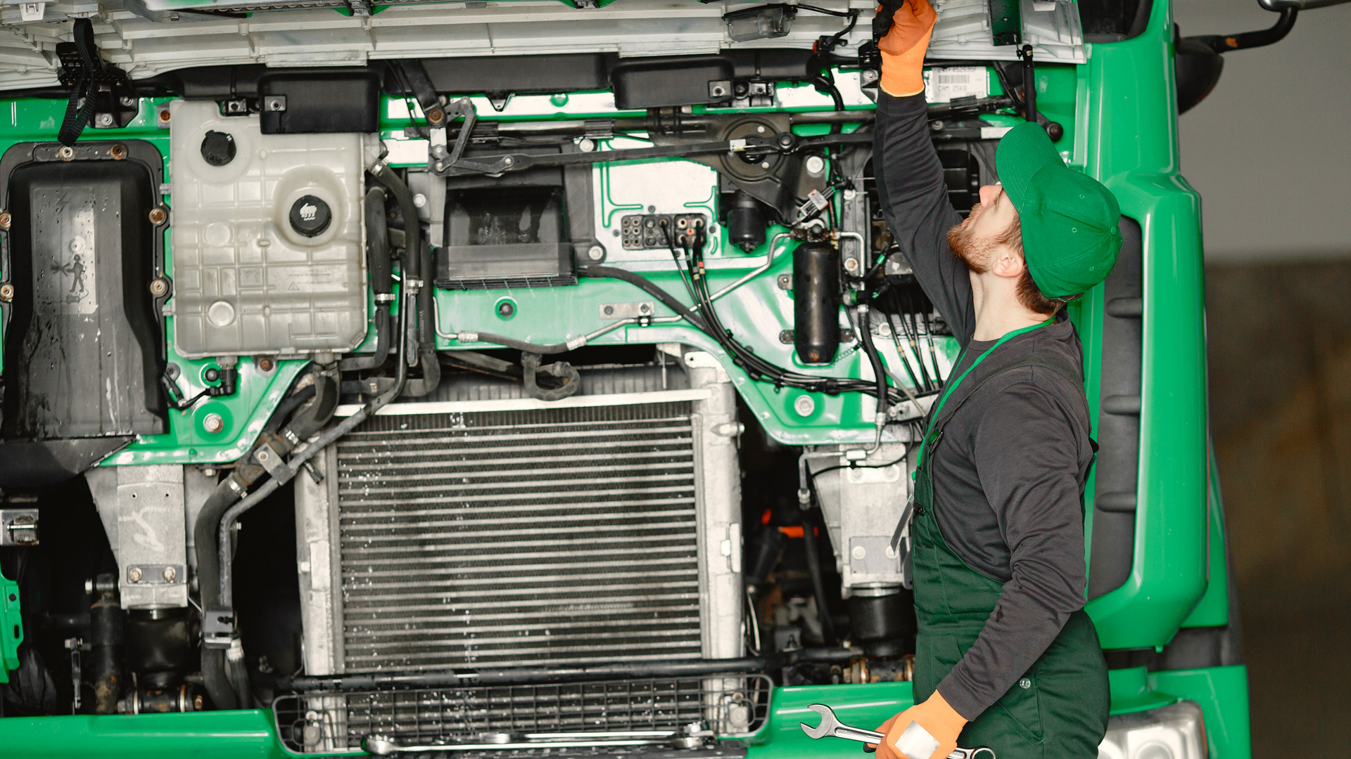 A mechanic in a green uniform, representing the leading fleet solutions provider, works on the engine of a large green truck, reaching inside the hood to make precise adjustments.