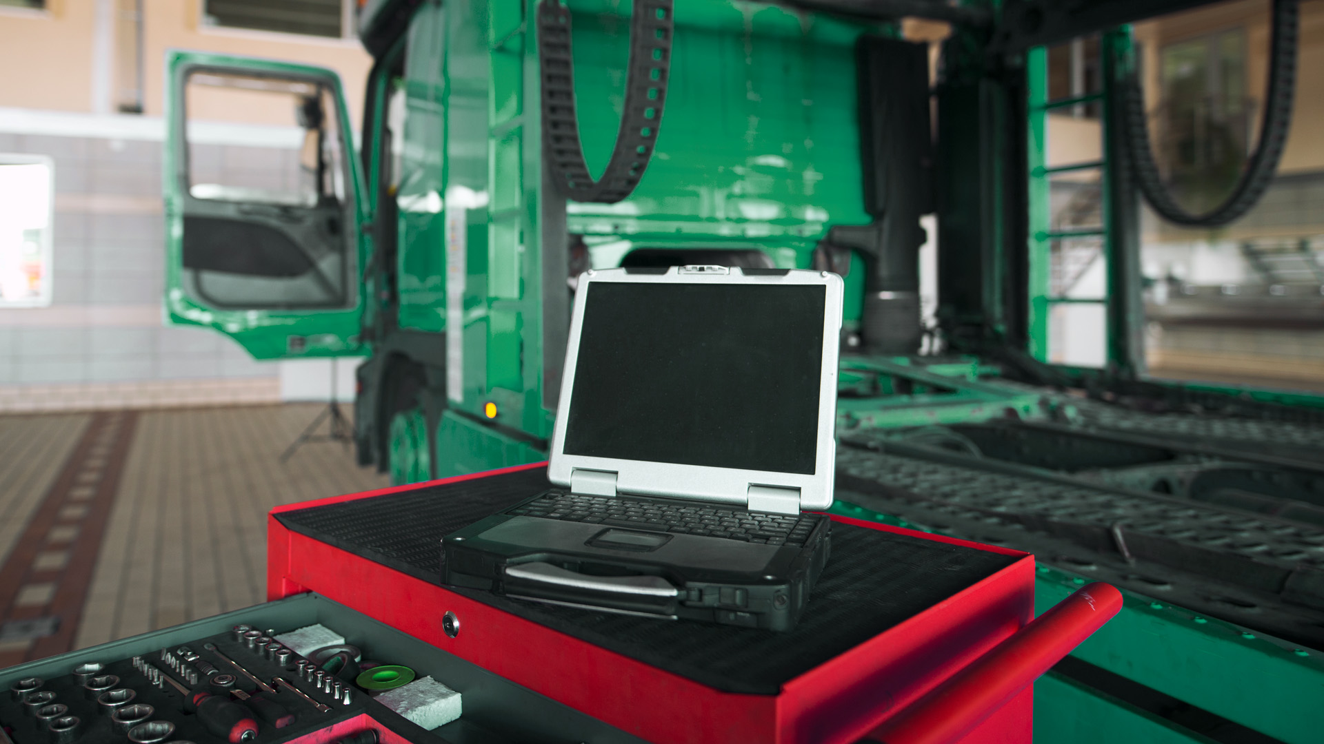 A diagnostic laptop from the leading fleet solutions provider sits on a red workstation in front of a green truck undergoing maintenance in a service garage.