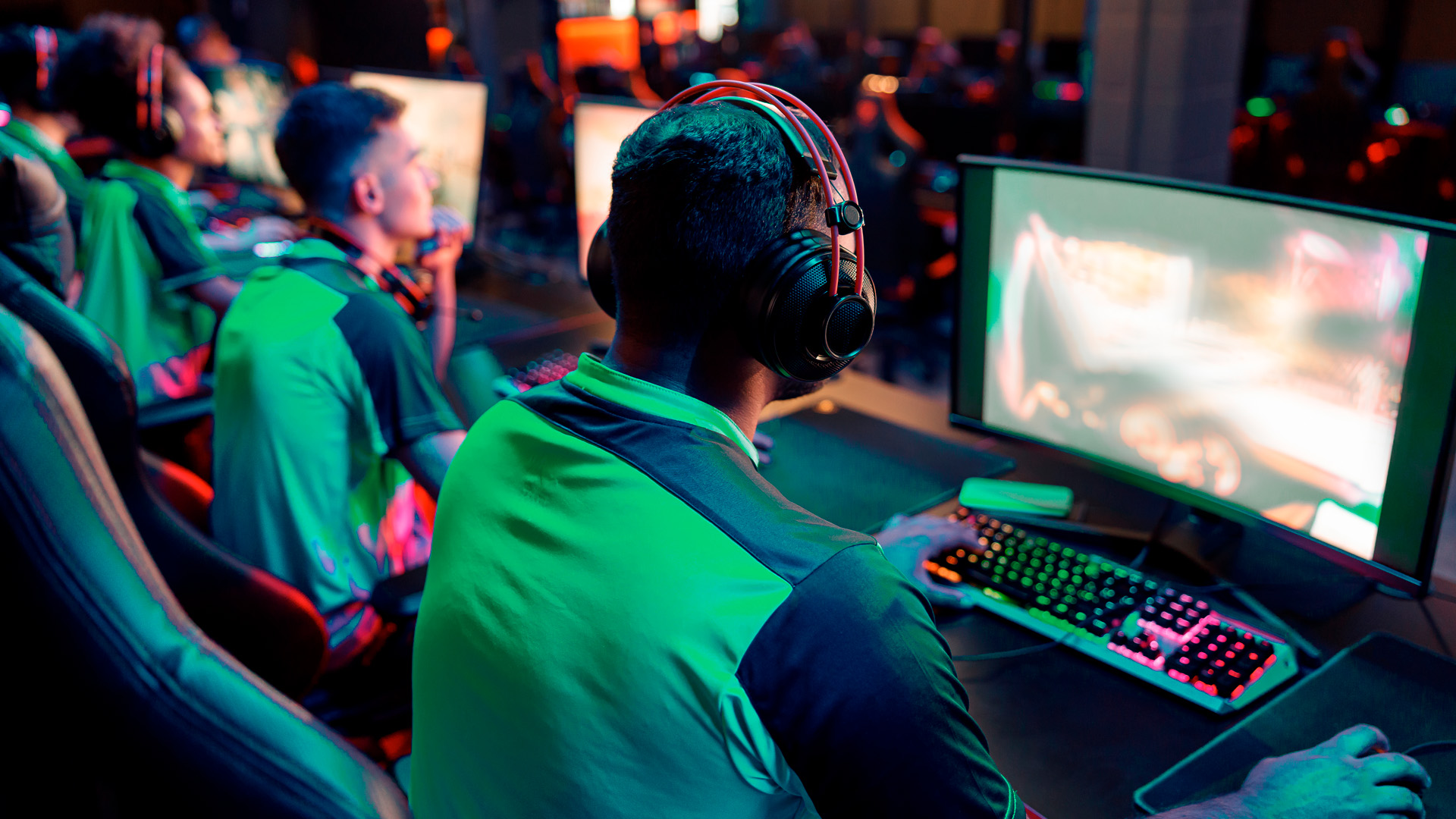 Gamers in Helix eSports jerseys wearing headphones and sitting at computer stations play in a dimly lit room with neon lights, focused on their screens.