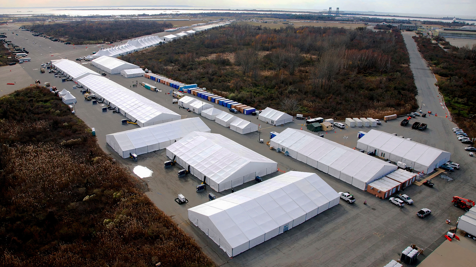 Aerial view of a temporary large-scale facility, featuring deployed resources like numerous white tents, trucks, and trailers arranged in rows on an open lot, surrounded by trees and located near an industrial area.