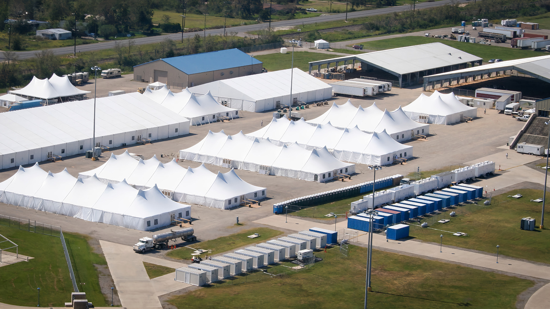 Aerial view of a temporary facility with large white tents and portable buildings, likely set up for emergency or event operations. Deployed resources are evident in the surrounding area, which includes roads, green spaces, and additional structures.