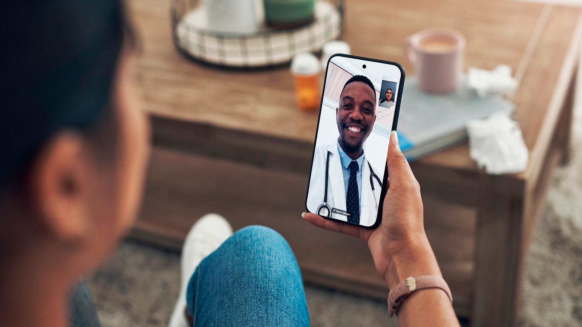 A person is using a smartphone, relying on Healthcare Solutions, to have a video call with a doctor. The doctor is smiling and wearing a white coat and stethoscope. A coffee table with items is visible in the background.