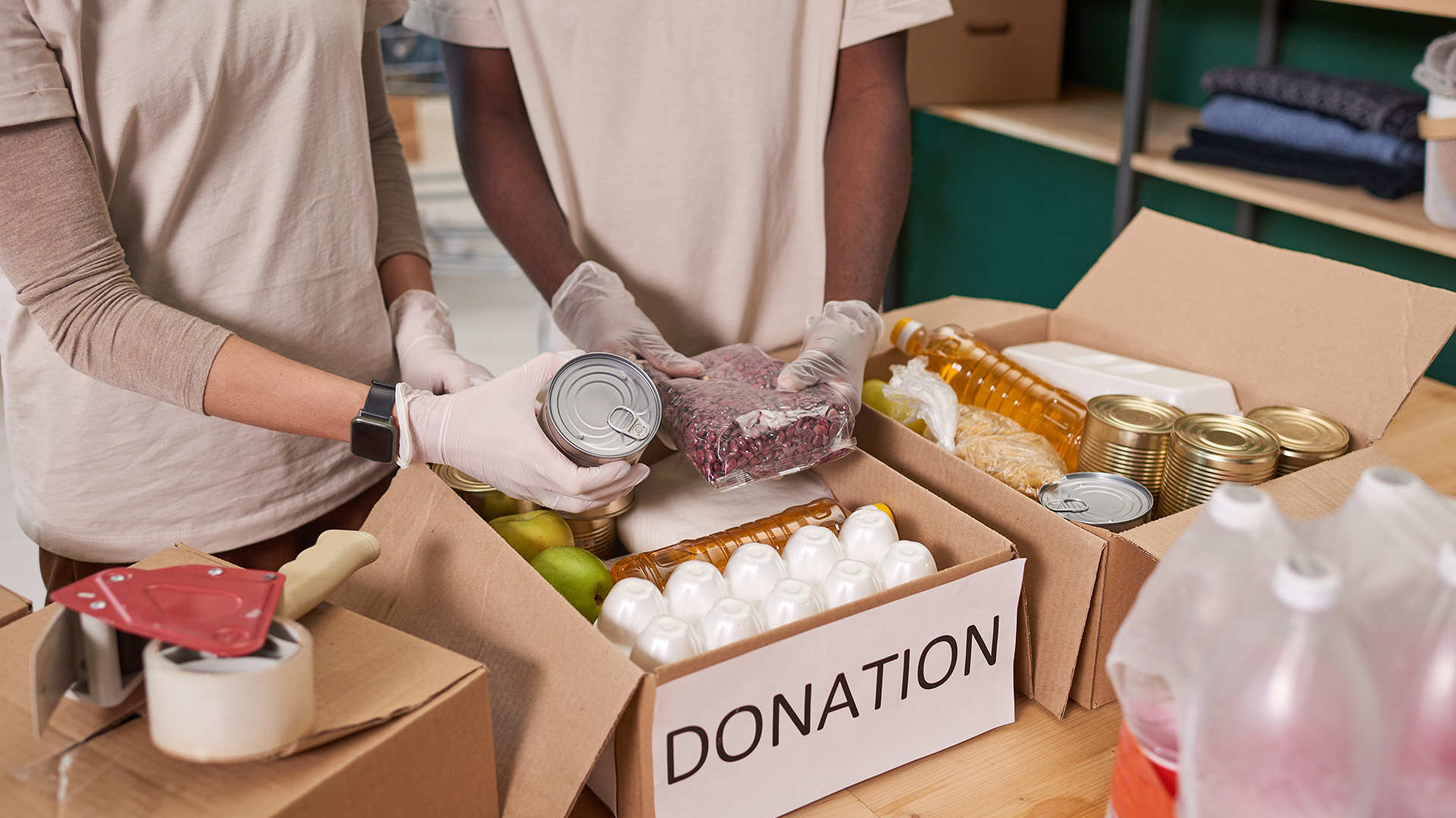 Two people packing food items into donation boxes, which include canned goods, eggs, and grains. They are wearing gloves and surrounded by other packing materials. As part of a global nonprofit initiative, their efforts are data-driven to efficiently maximize the impact of donations.