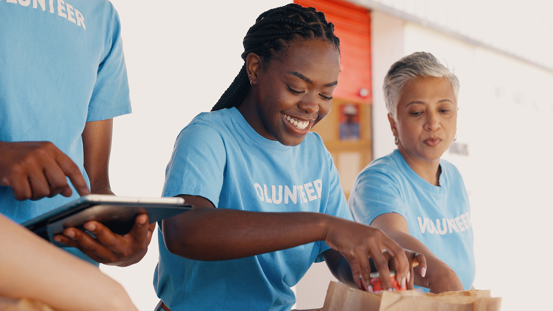 Three volunteers in light blue shirts pack items into brown paper bags. One handles a tablet, focusing on data management while the others work together and smile. They are inside a well-lit area with a red door in the background, embodying the spirit of a global nonprofit organization.