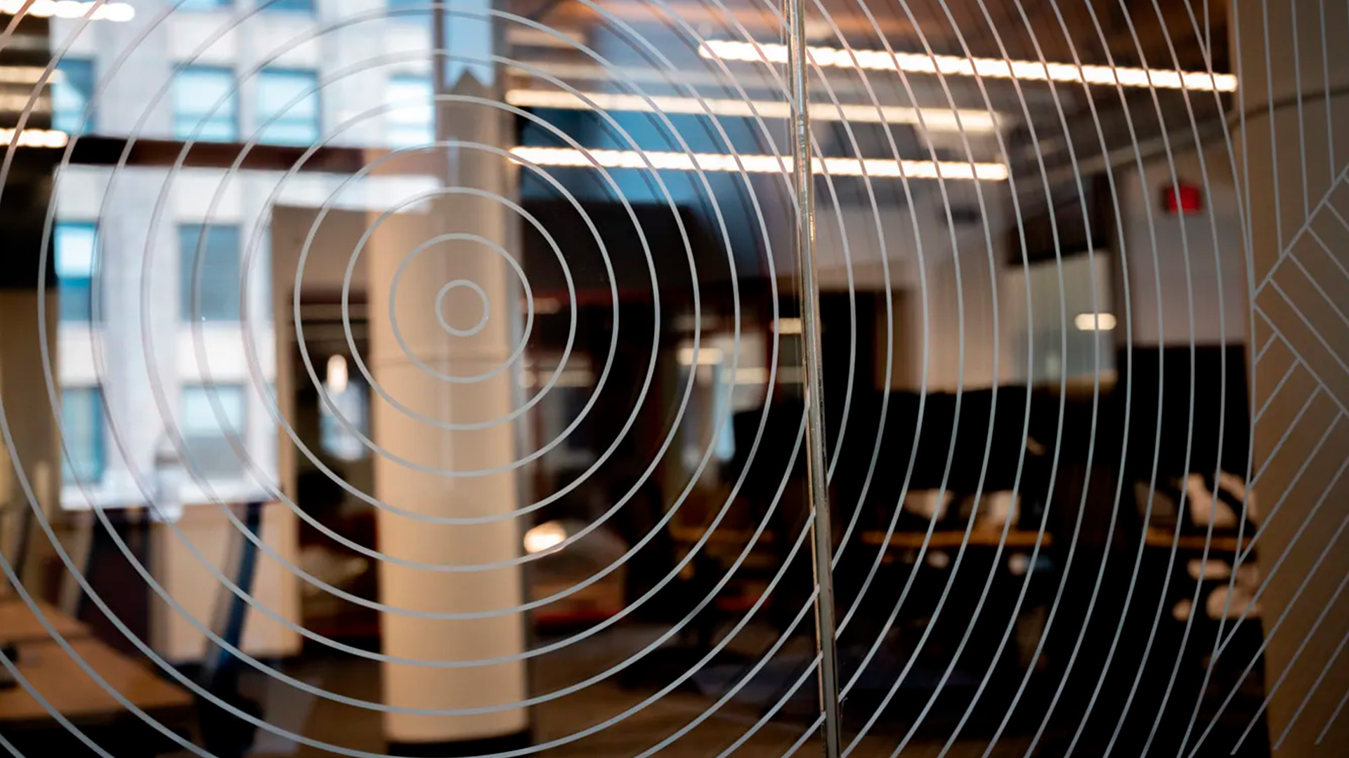 Close-up of a Kanda glass partition with concentric circle designs in a modern office space, featuring desks, chairs, and overhead lighting.